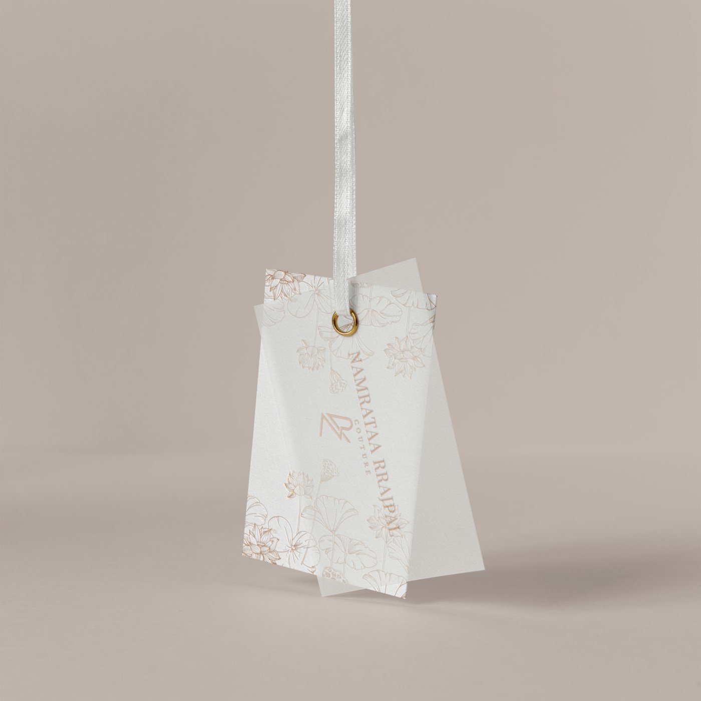 Namrataa Rrajpal Couture cloth tag with a gold grommet and lotus line art, shot hanging against a warm neutral backdrop
